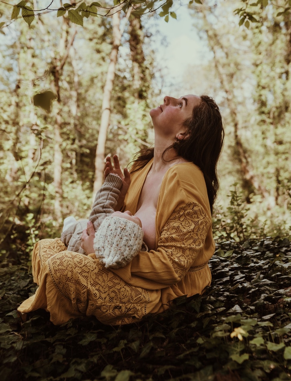 Barbora breastfeeding her baby in the woods, looking up at the trees